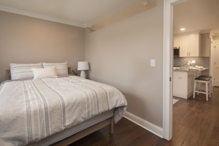 The Courtyard @ CliftonA cozy bedroom with a neatly made bed and view into a modern kitchen with barstools.