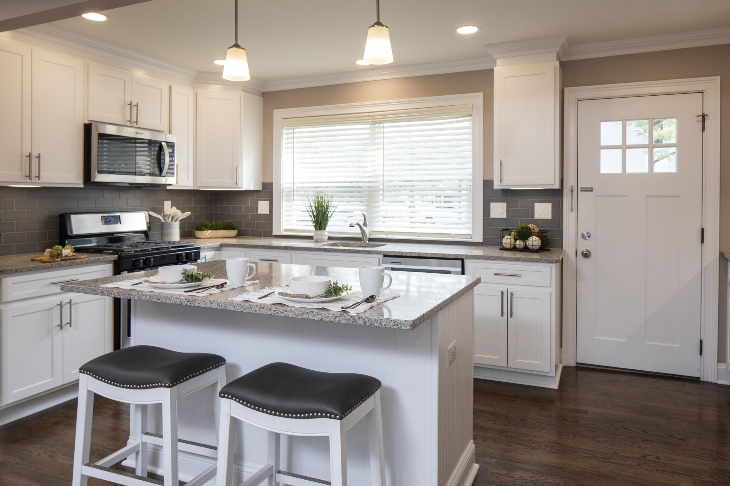 Modern kitchen with white cabinets, an island with two stools, and sunlight streaming through a large window.