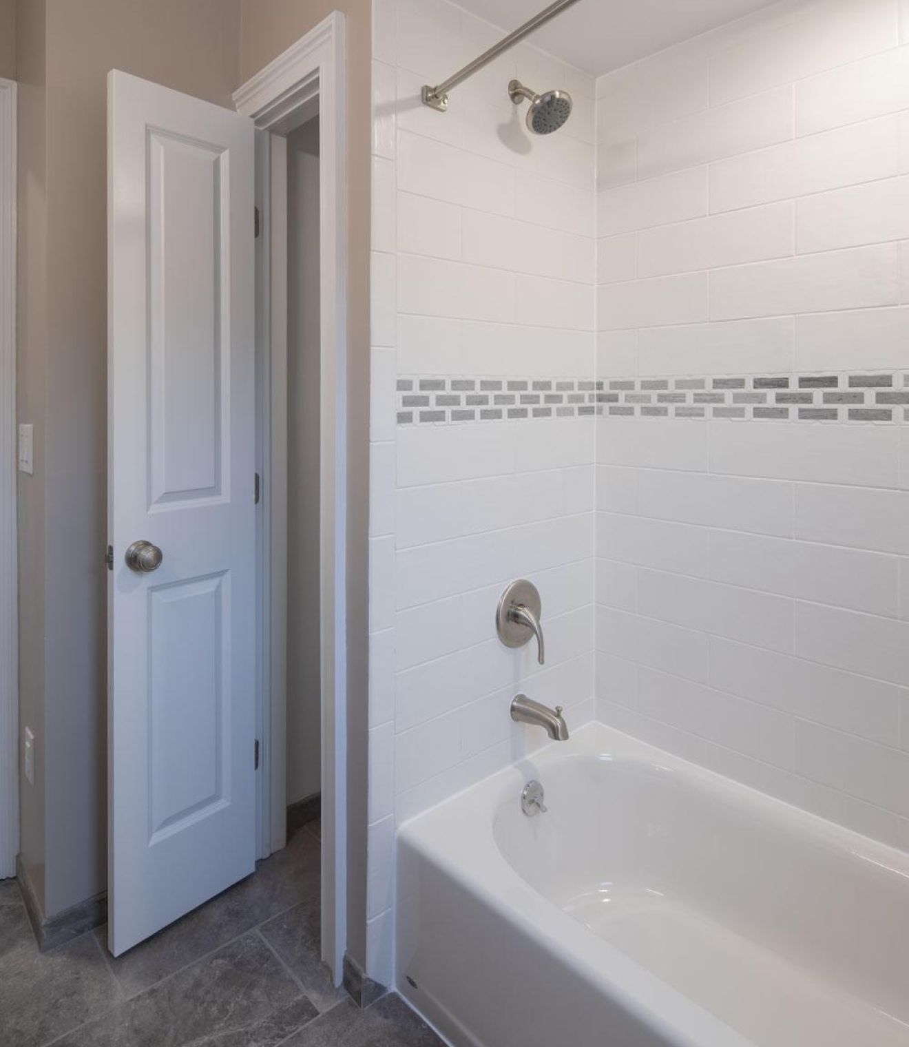 Modern bathroom with white tile bathtub, gray accent strip, chrome fixtures, and open door to hallway.