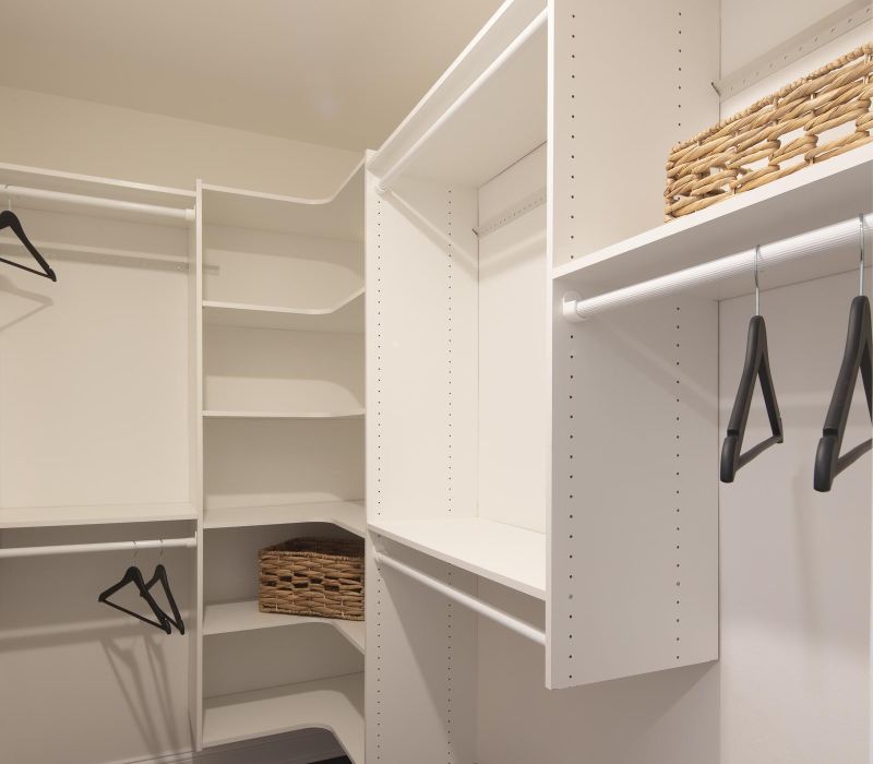 White walk-in closet with shelves, hanging rods, black hangers, and wicker baskets on dark wood floor.