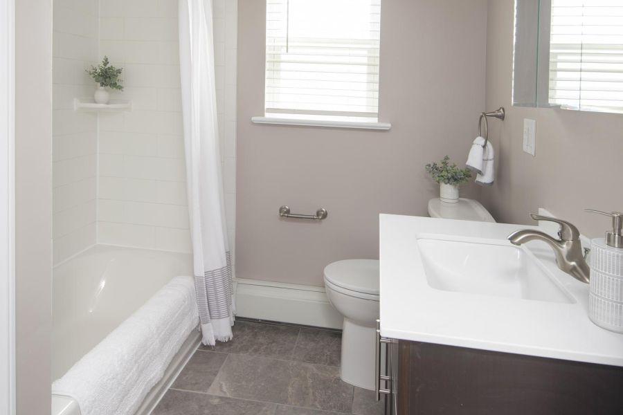 The Courtyard @ Clifton Modern bathroom with gray tile floor, white bathtub and shower, vanity, toilet, and two small windows.