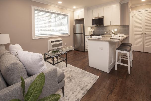 Modern studio apartment with a small kitchen, gray sofa, and natural light from a large window.