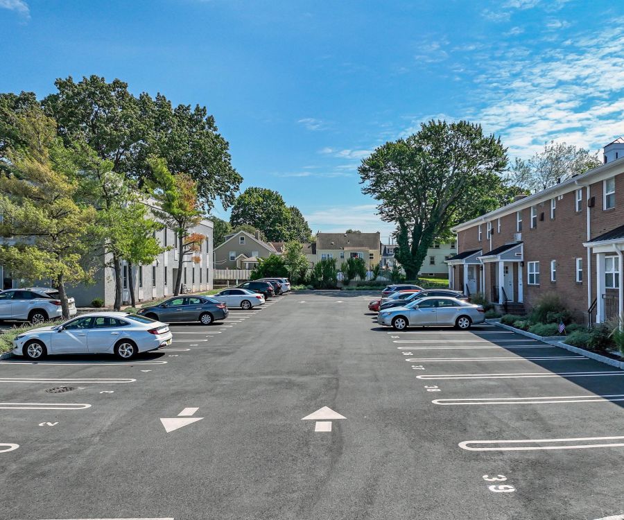 Parking lot with cars parked on both sides, surrounded by buildings and trees under a blue sky.