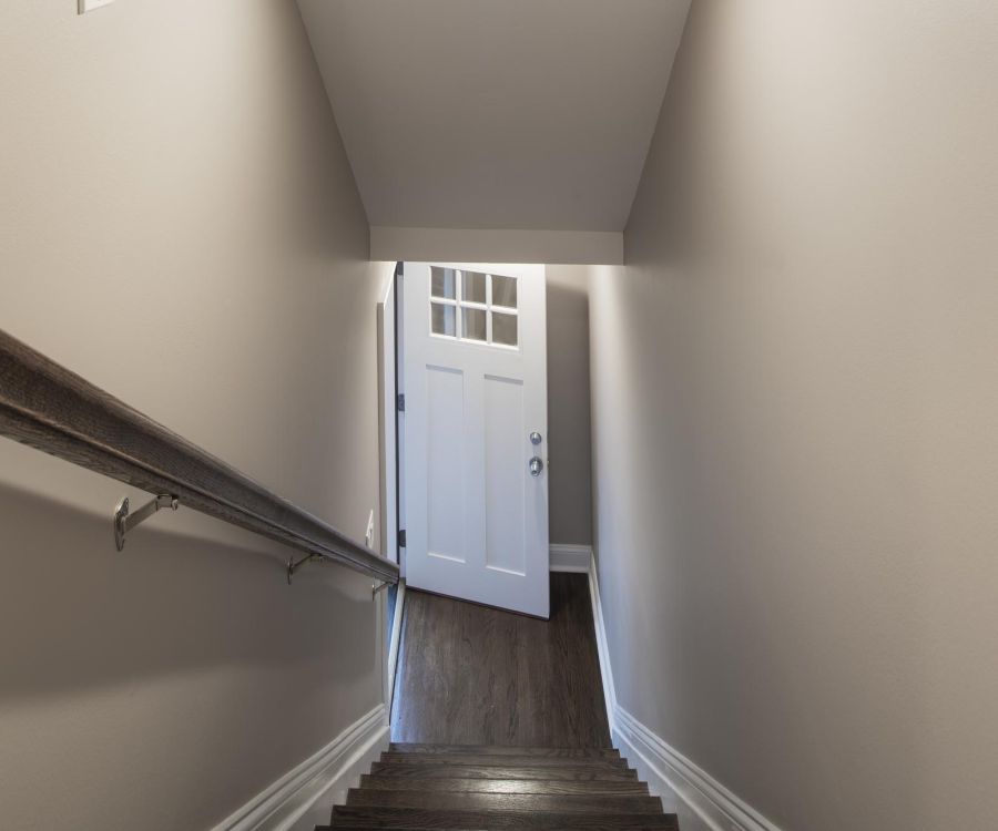 Wooden stairs leading down to a partially open white door in a narrow hallway with beige walls.