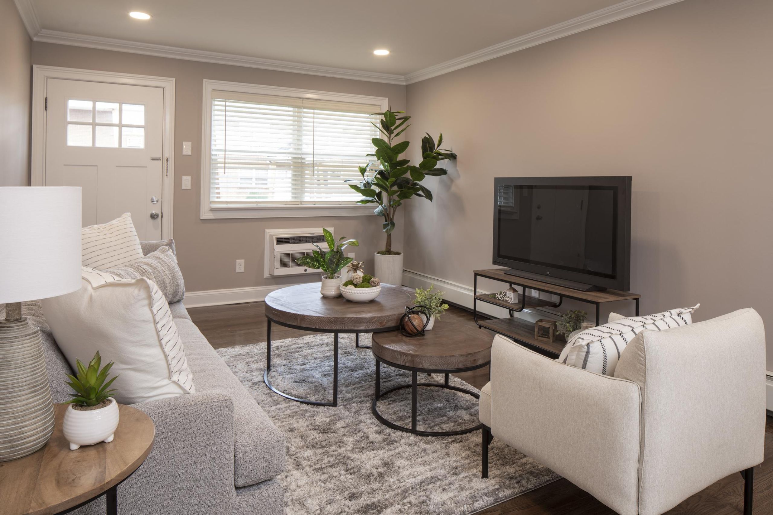 The Courtyard @ CliftonBright living room with beige walls, a sofa, coffee table, TV, armchair, and large window with blinds.