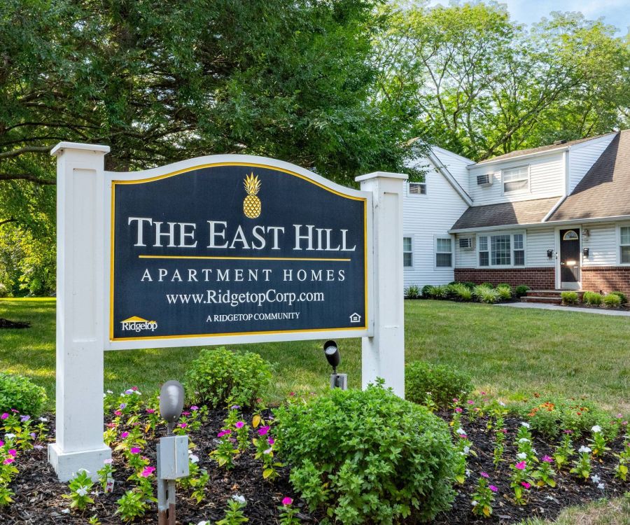 A sign for The East Hill Apartment Homes stands in front of a white house and landscaped garden.