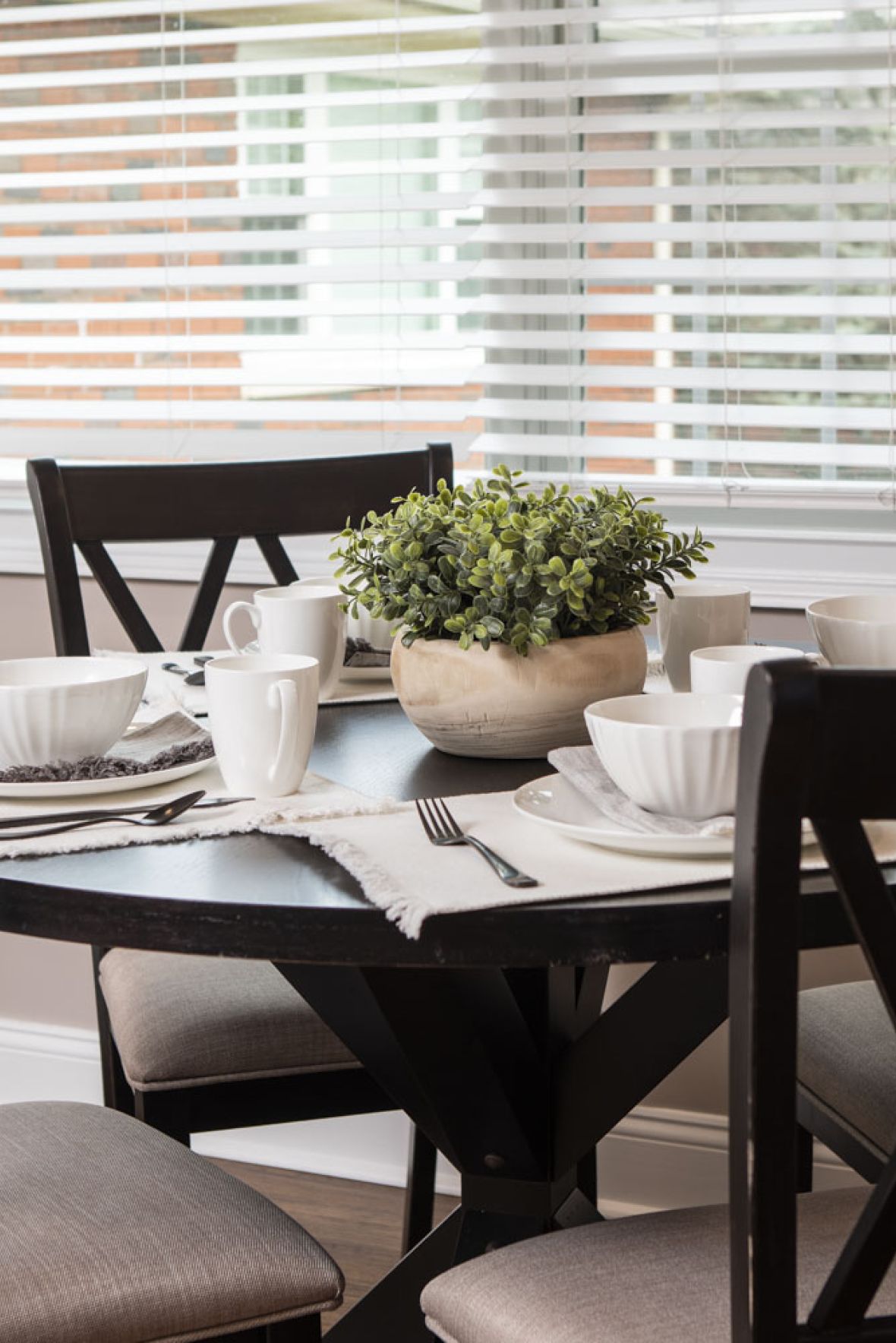 The East Hill A round dining table set for four with white dishes and a potted plant centerpiece near a window with blinds.