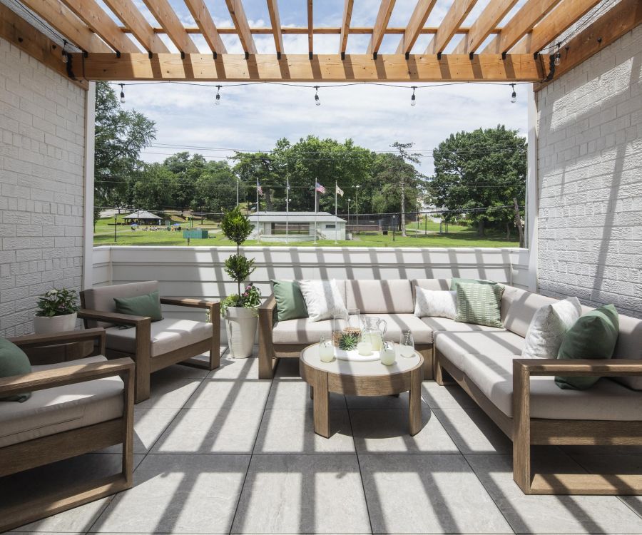 Patio with wooden pergola, outdoor seating, chairs, and plants, overlooking a green lawn with flags.