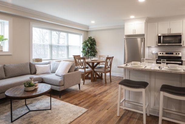 Modern open-concept kitchen and living room with neutral decor, wood floors, and natural light from large windows.
