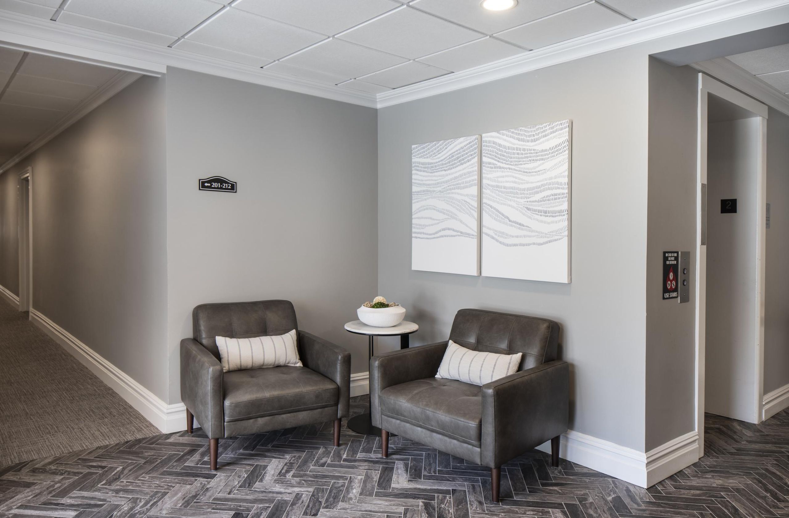 The Parkview @ Polifly Two gray armchairs with pillows sit by a small table in a modern, gray-toned hallway near an elevator.