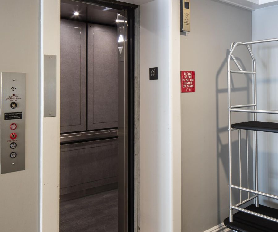 An open elevator with dark interior next to a luggage cart in a hallway with gray herringbone flooring.