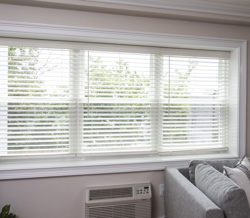 Large window with white blinds partially open, next to a gray sofa and air conditioner in a bright room.