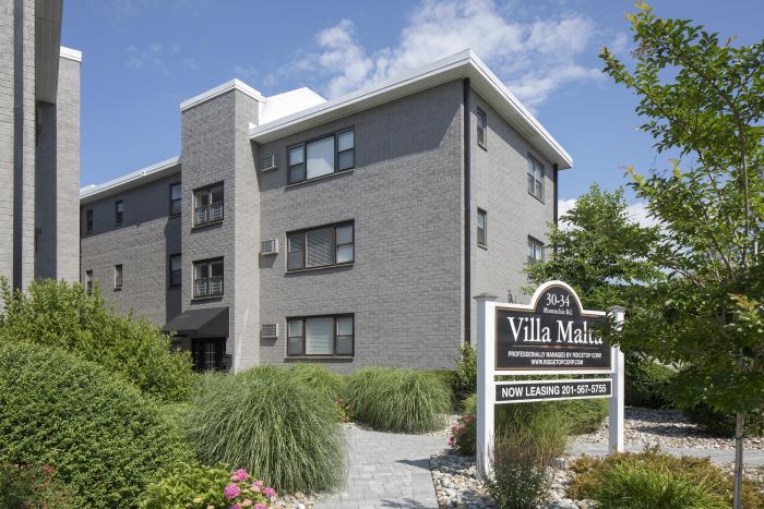 Villa Malta Gray brick apartment building with a Villa Malta leasing sign, surrounded by greenery and blue sky.