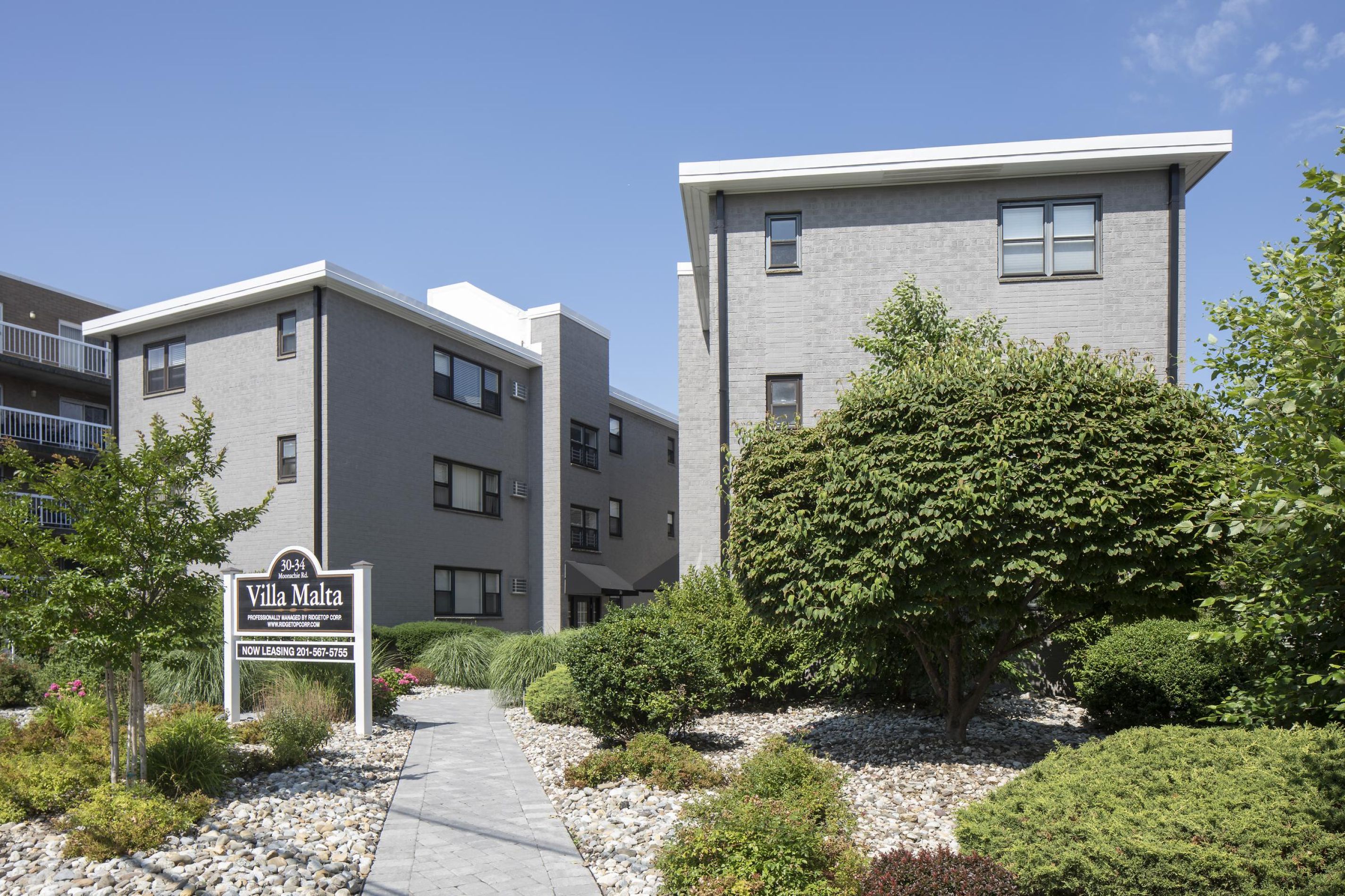 Gray apartment buildings with a "Villa Malta" sign, surrounded by landscaped bushes and a stone walkway.