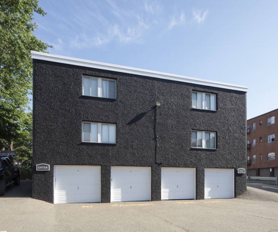 A black stucco apartment building with four white garage doors and parked cars on a clear day.
