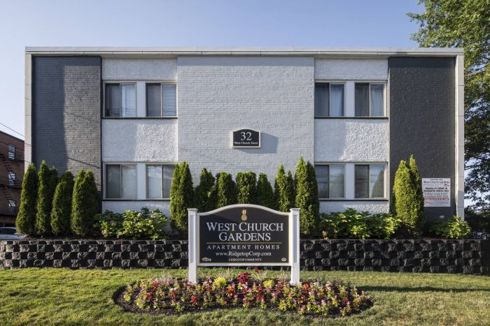 A gray apartment building with a "West Church Gardens Apartment Homes" sign and neatly trimmed bushes in front.