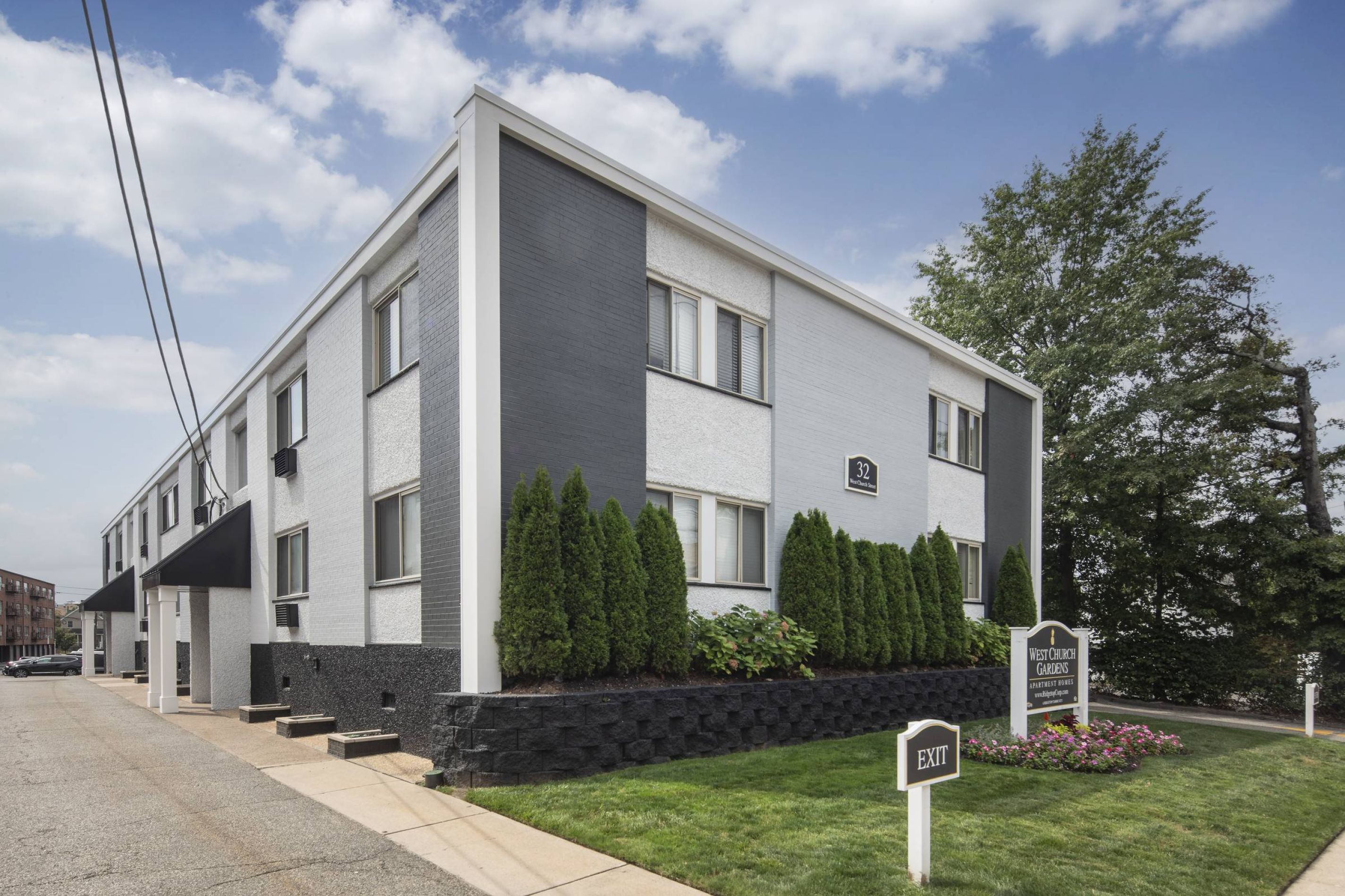 Two-story modern apartment building with gray and white exterior, shrubs, and a sign reading "Riverton Gardens.