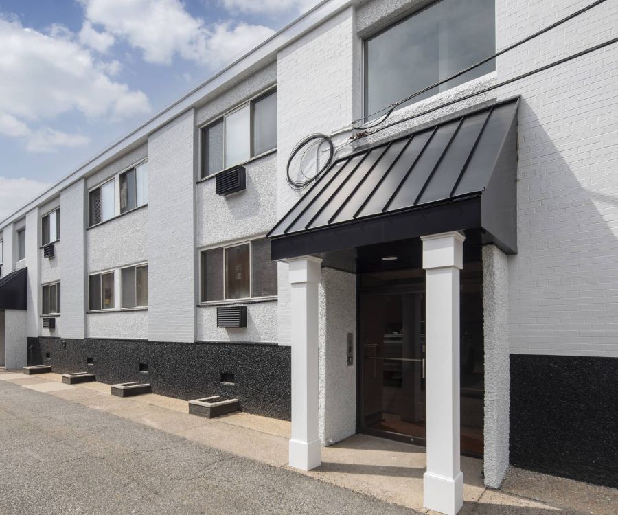 Two-story white brick building with black awning, large windows, and a paved walkway under a partly cloudy sky.