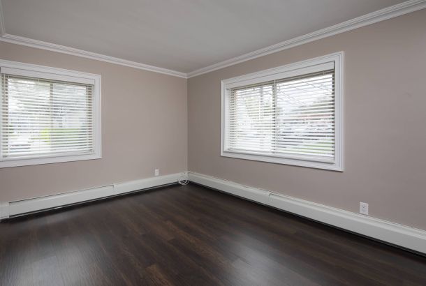 Empty room with dark wood floors, beige walls, white trim, and two large windows with white blinds.