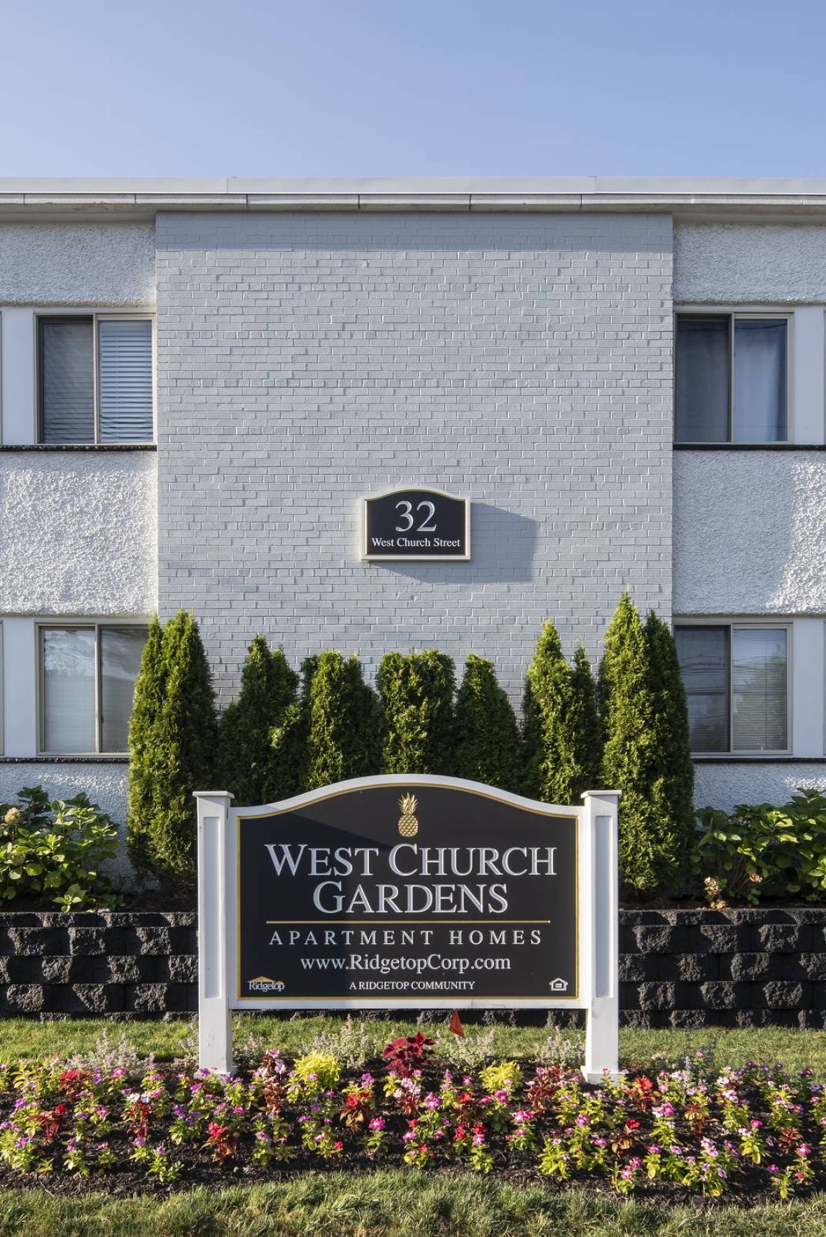 West Church Gardens Empty outdoor tennis court with nets, surrounded by trees and buildings, under a partly cloudy sky.