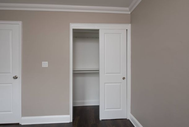 Empty closet with white sliding doors in a room with taupe walls and dark wood flooring.