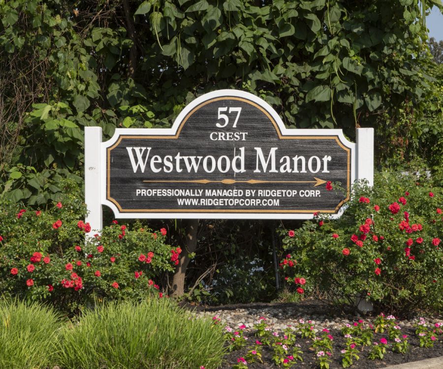 A wooden sign reading "57 Crest Westwood Manor" with flowers and greenery in the background.