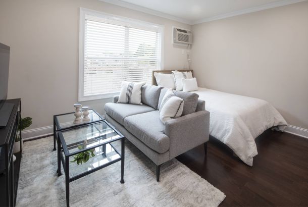 A small, bright bedroom with a bed, gray sofa, glass coffee table, rug, and window with blinds.