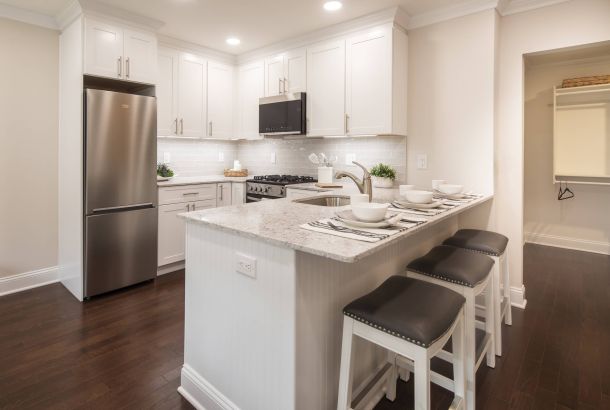 Modern white kitchen with stainless steel appliances, marble island, and three bar stools set for dining.