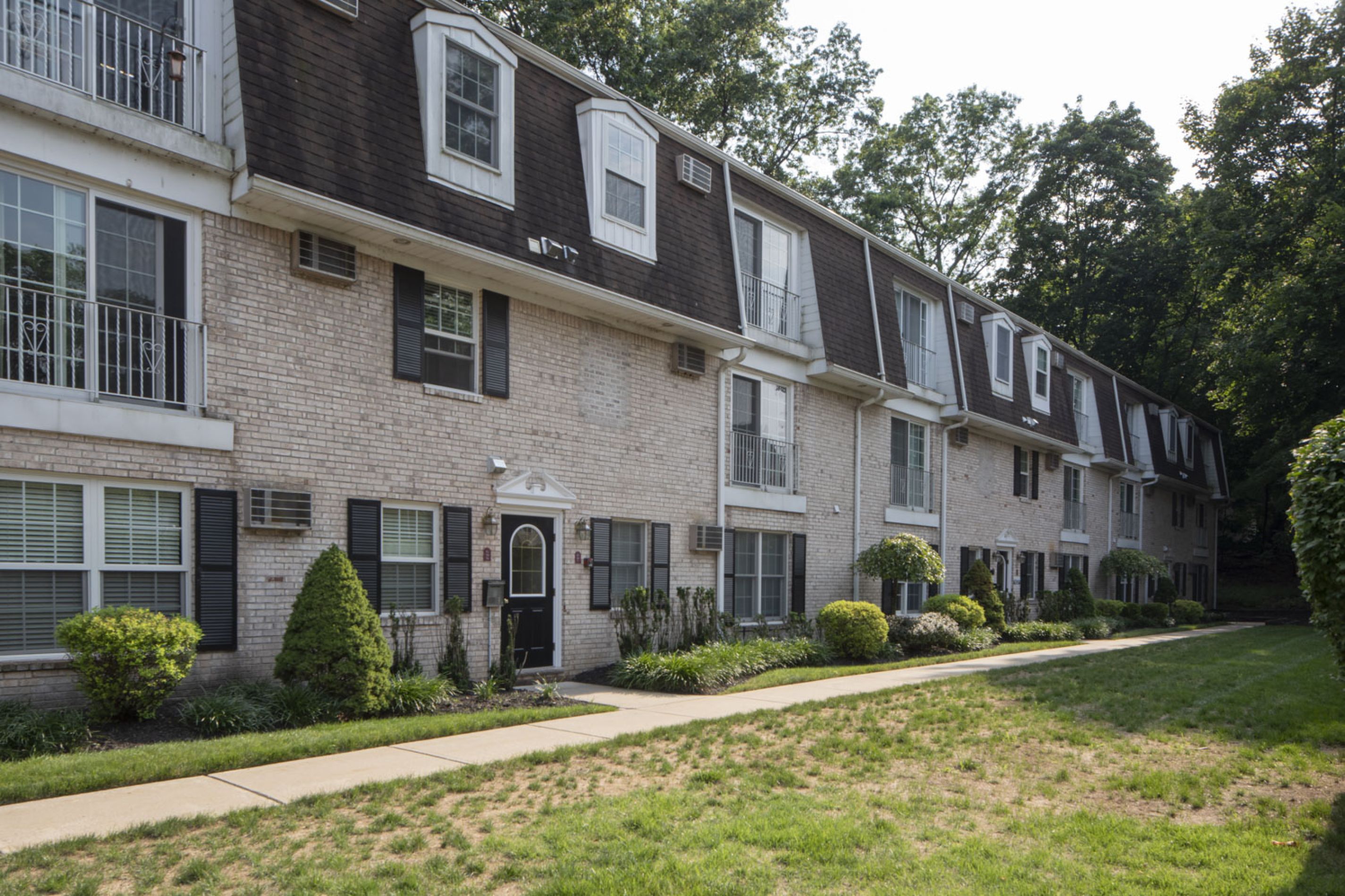 A row of beige brick townhouses with black doors, dormer windows, and small shrubs in front.