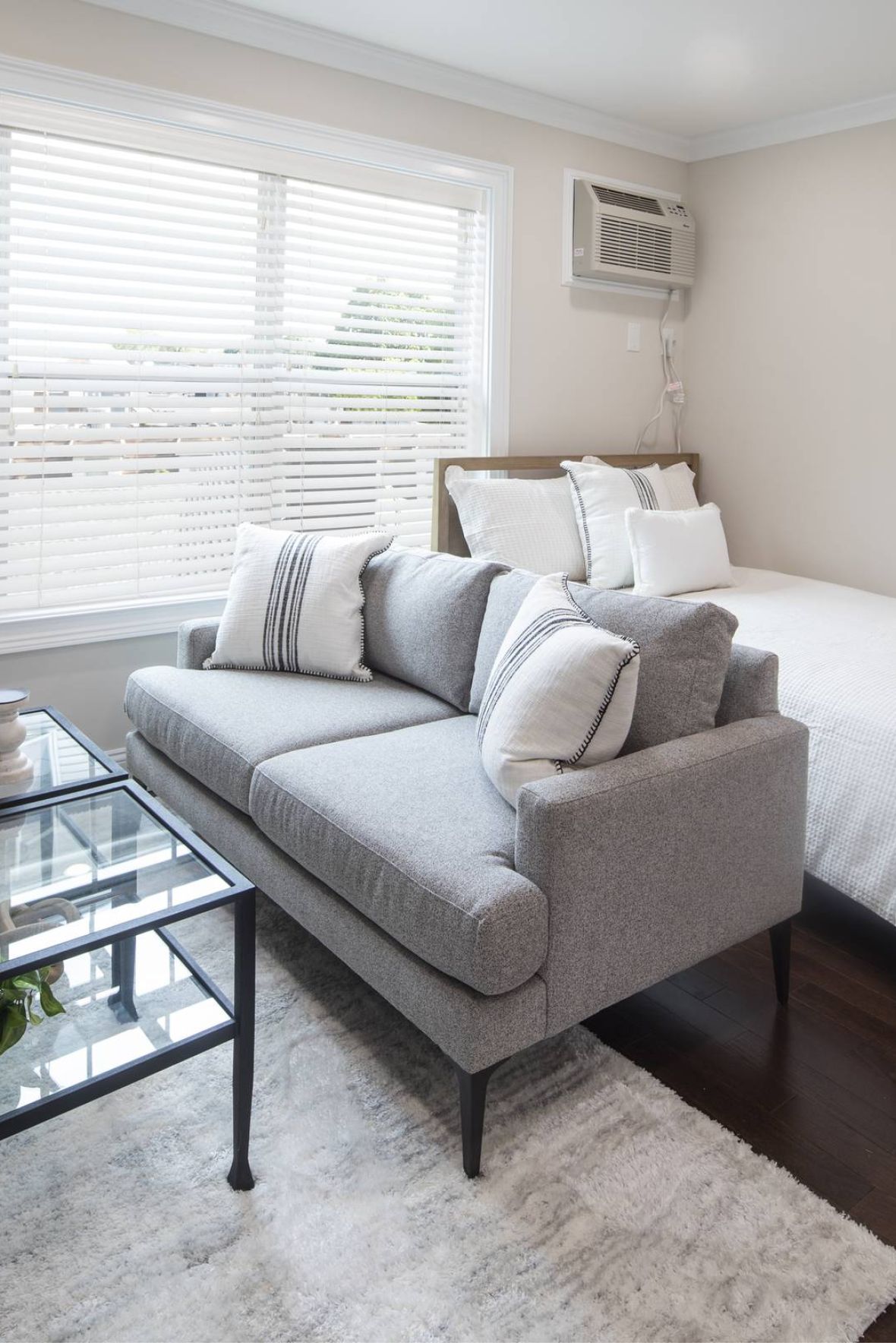 A small, bright bedroom with a bed, gray sofa, glass coffee table, rug, and window with blinds.