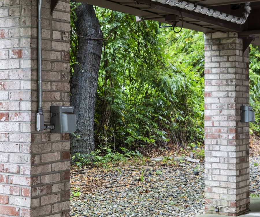 Three brick columns with electrical boxes stand under a roof, with trees and rocks in the background.