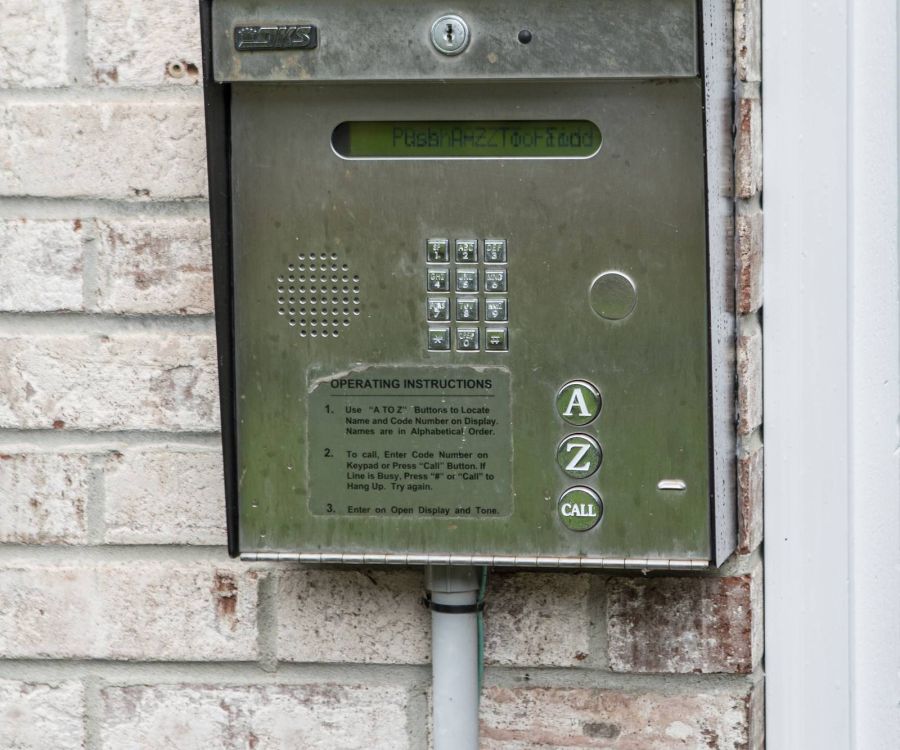 A metal keypad entry system mounted on a brick wall, with buttons and a small display screen.