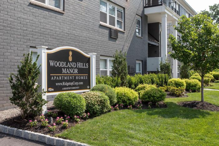Sign for Woodland Hills Manor Apartment Homes in front of a gray brick building with landscaped bushes and grass.
