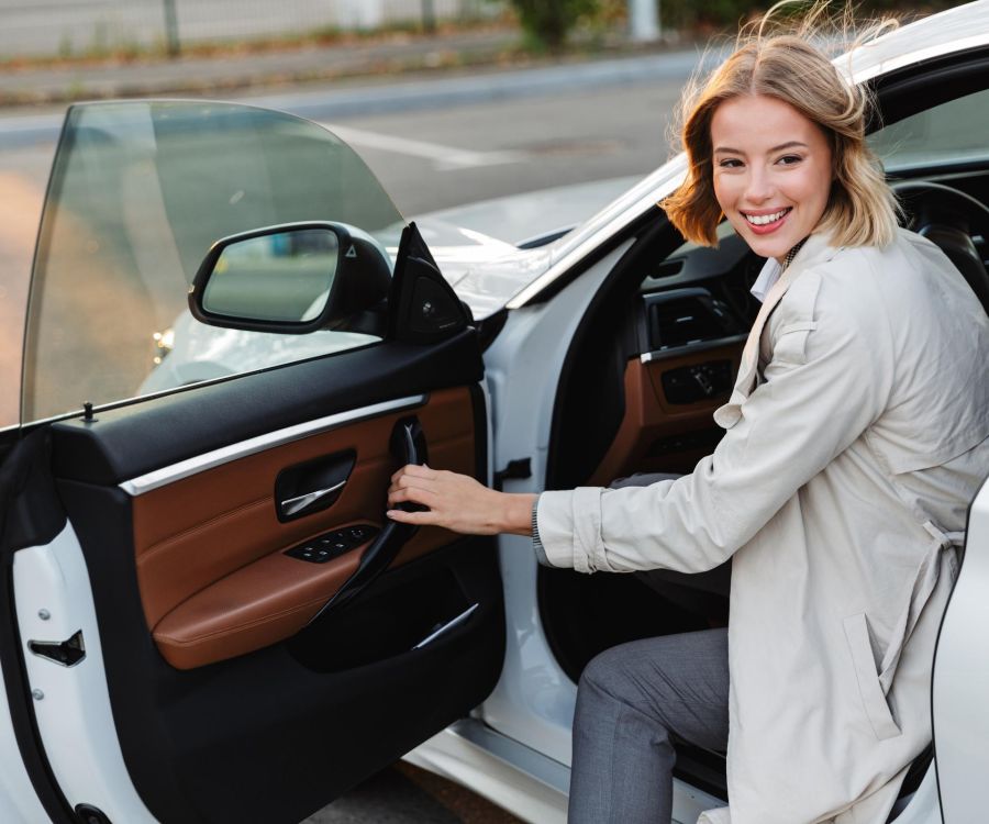 Smiling woman in a light coat sits in a car with the door open, looking back at the camera.