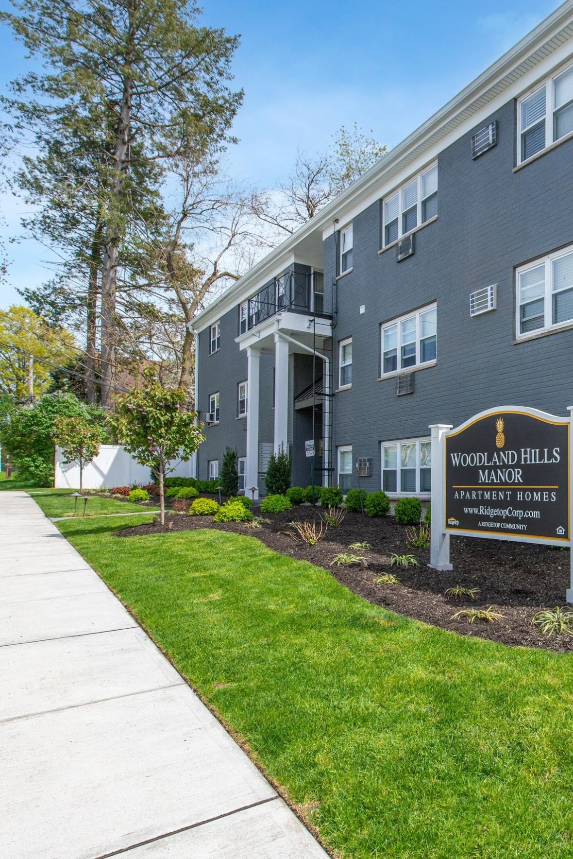 Woodland Hills Manor Empty outdoor tennis court with nets, surrounded by trees and buildings, under a partly cloudy sky.