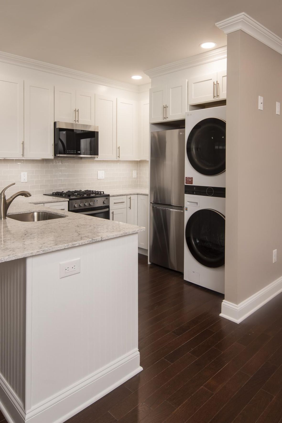 Modern kitchen with white cabinets, marble countertop, stacked washer and dryer, and dark wood flooring.