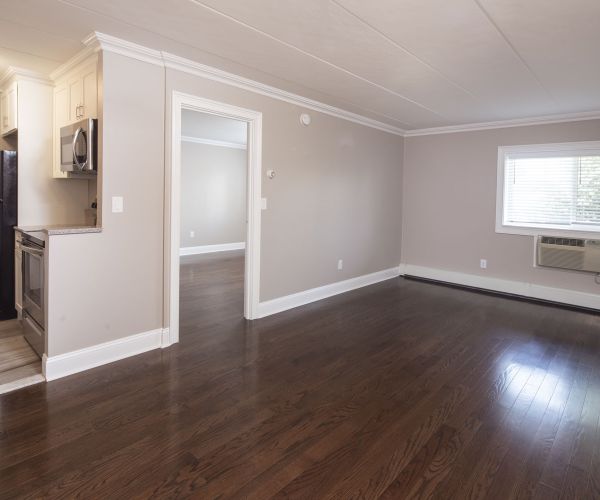 Empty Cadillac Manor apartment with hardwood floors, a small kitchen, white walls, and a window with an air conditioning unit.