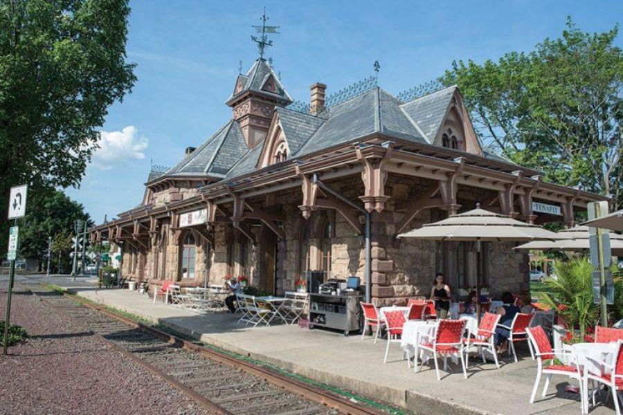 Historic stone train station in Tenafly with outdoor café seating beside train tracks on a sunny day.