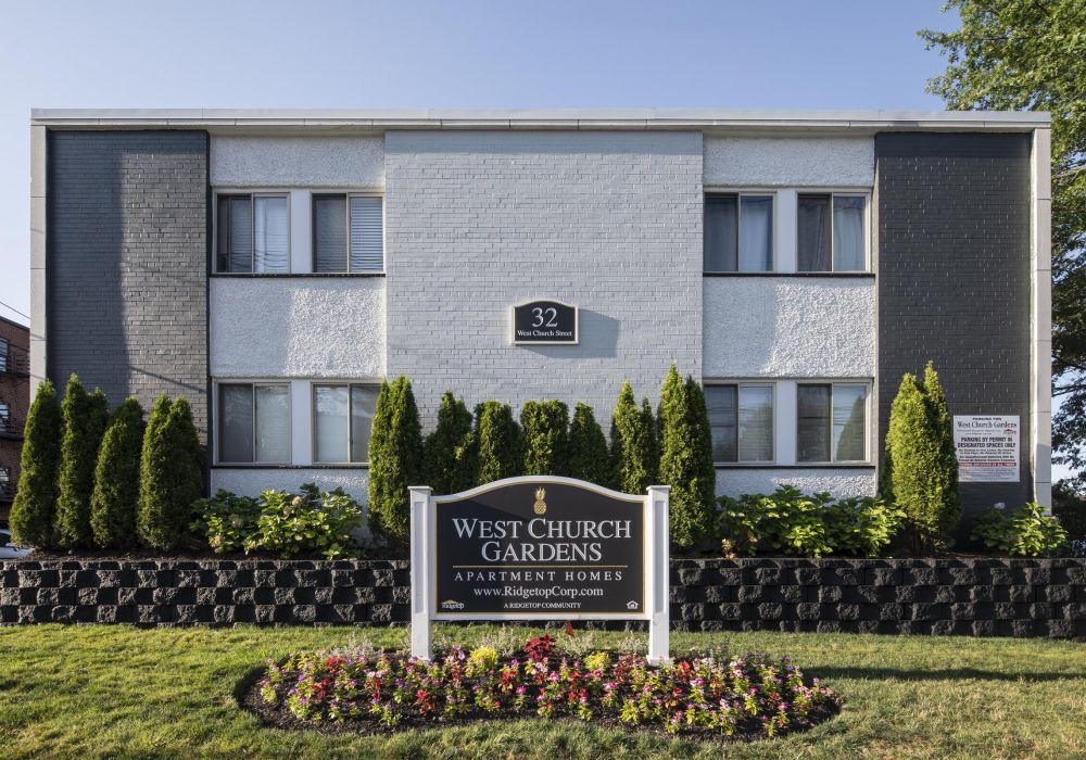 A gray and white apartment building with a "West Church Gardens" sign in front, welcoming you home.