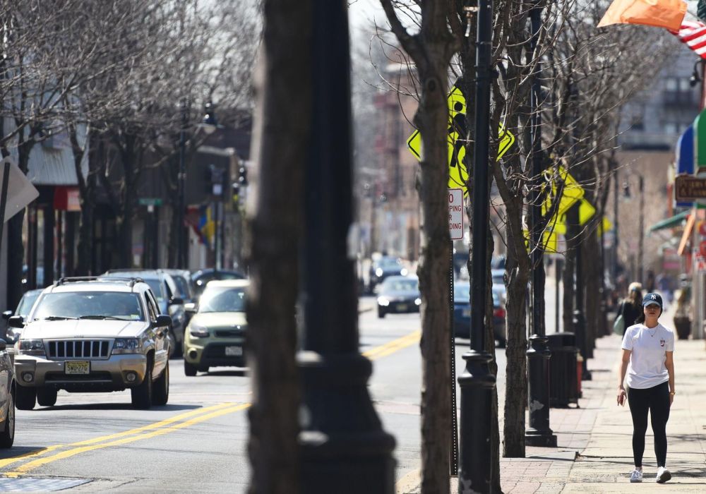 A woman walks on a Bergenfield city sidewalk beside parked cars and traffic on a sunny day.