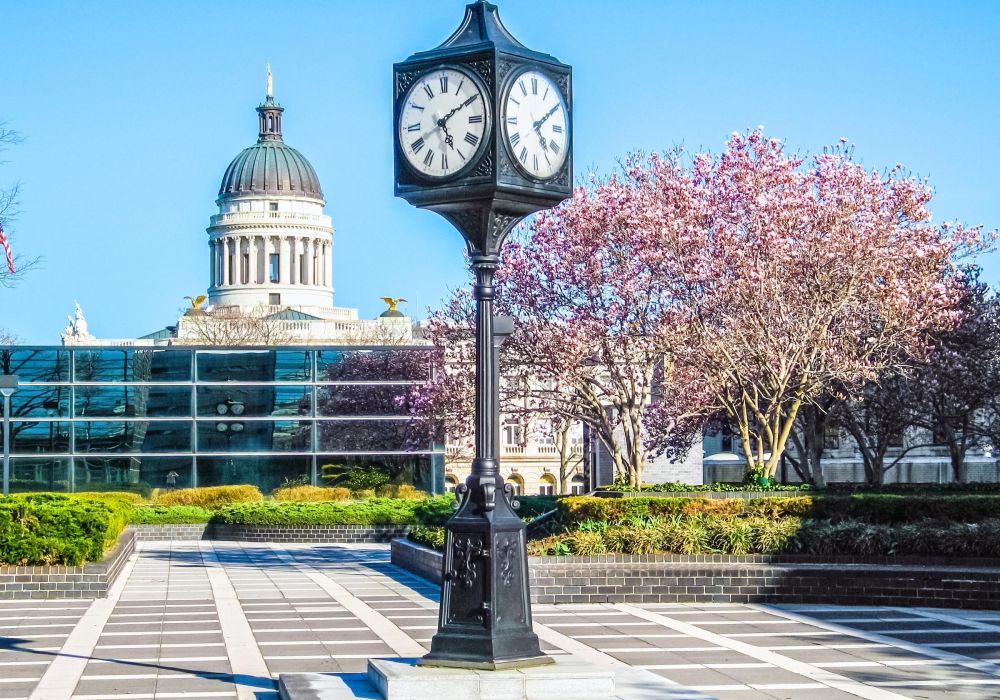 A clock stands in a plaza with blooming trees and a domed building in the background under a clear sky.