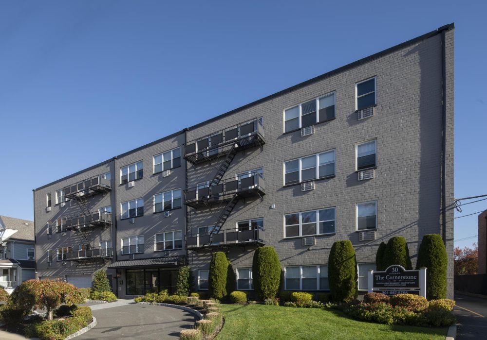 Four-story gray apartment building with balconies and landscaping under a clear blue sky.