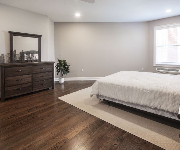 Minimalist bedroom with wooden floor, white bedding, dresser, nightstand, and large window letting in natural light.