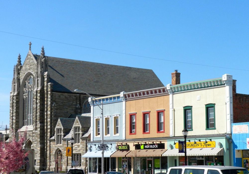 Colorful storefronts in Leonia and a large stone church under a clear blue sky with cars parked in front.