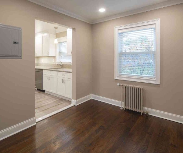 Empty room with dark wood floor, beige walls, radiator, window, and a view into a white kitchen.