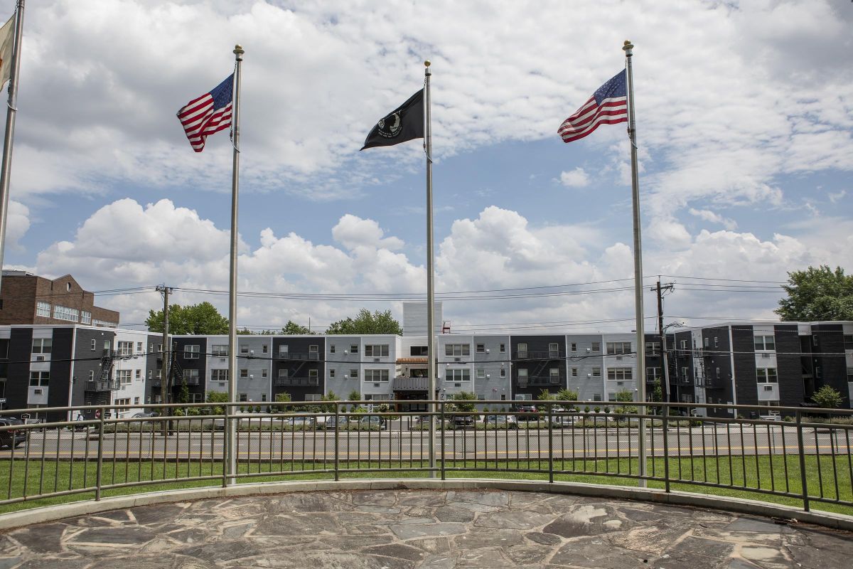 Three flags on poles—two American, one black—fly in front of modern apartment buildings under a partly cloudy sky.