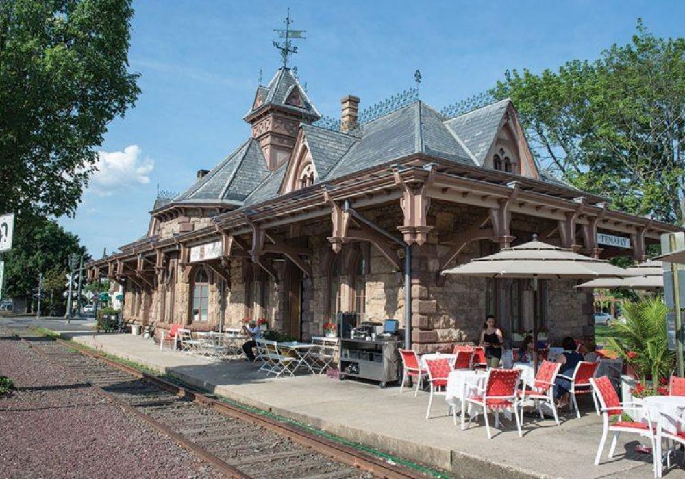 Historic stone train station in Tenafly with outdoor café seating beside train tracks on a sunny day.
