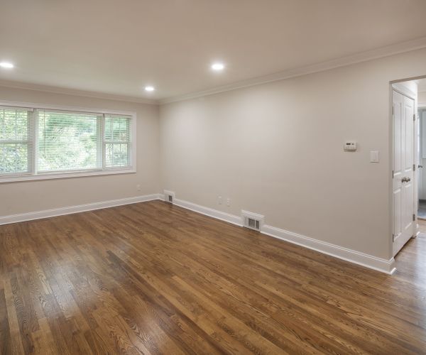 Bright, empty room with hardwood floors, large window, beige walls, and recessed ceiling lights.