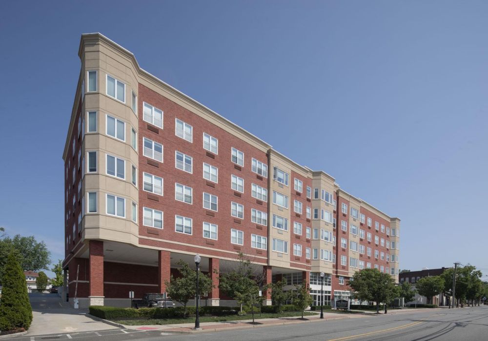 Five-story red and beige apartment building on a street corner, with trees and a clear blue sky.