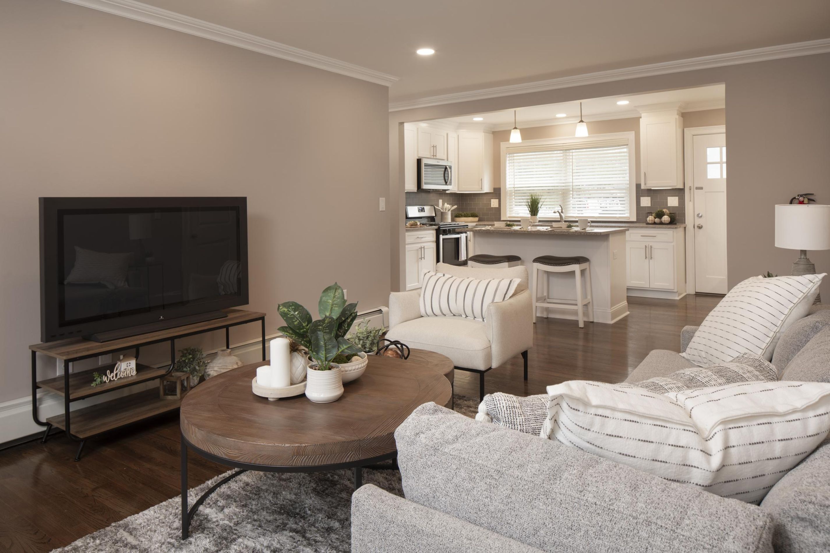 Modern living room with gray sofas, a round wooden coffee table, and view into a bright kitchen with barstools.